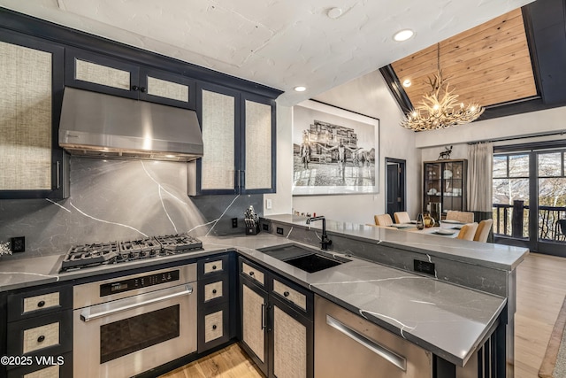 kitchen with an inviting chandelier, a peninsula, a sink, under cabinet range hood, and appliances with stainless steel finishes