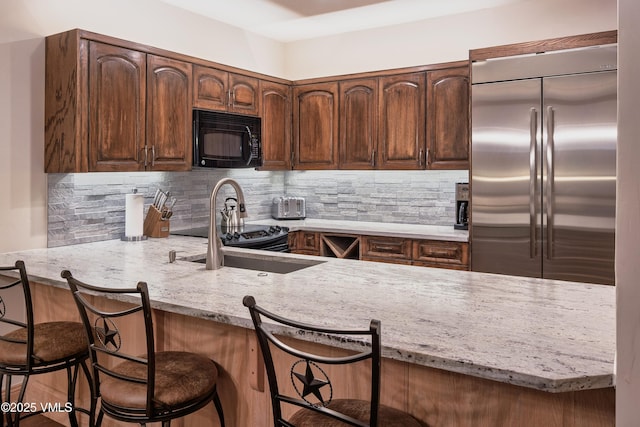 kitchen with decorative backsplash, light stone counters, black microwave, a sink, and built in fridge