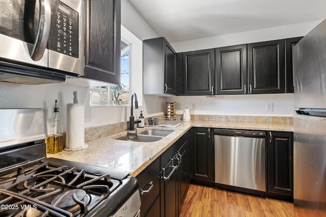 kitchen with dark cabinets, a sink, appliances with stainless steel finishes, light stone countertops, and light wood finished floors