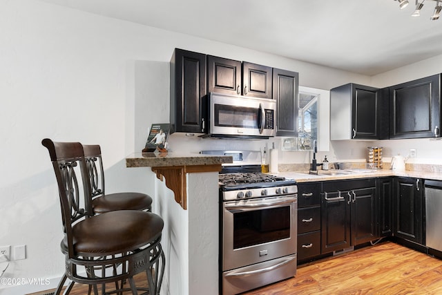 kitchen featuring light wood finished floors, appliances with stainless steel finishes, a sink, dark cabinetry, and a kitchen breakfast bar