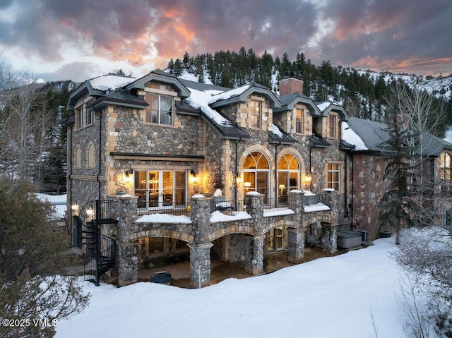 snow covered house featuring stairs, stone siding, and a chimney