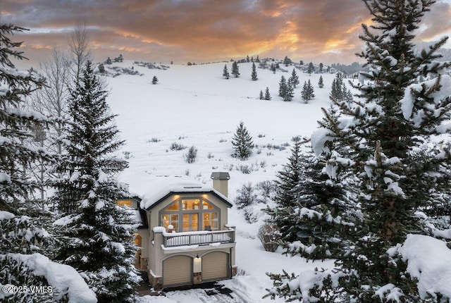snow covered rear of property featuring a chimney, a balcony, and a garage