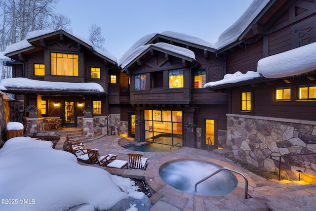 snow covered back of property featuring stone siding, a patio, and a hot tub