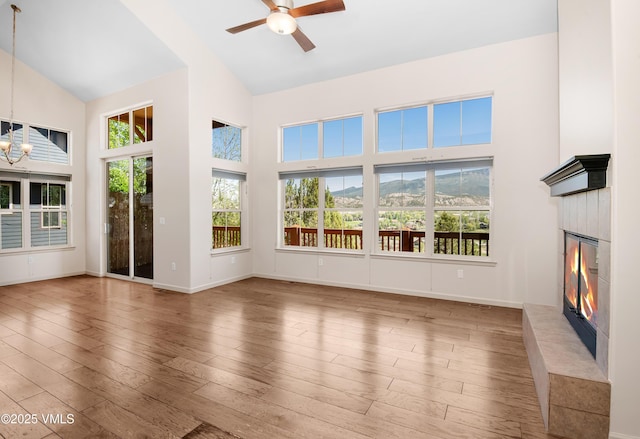 unfurnished living room with high vaulted ceiling, a tiled fireplace, a chandelier, a ceiling fan, and wood finished floors