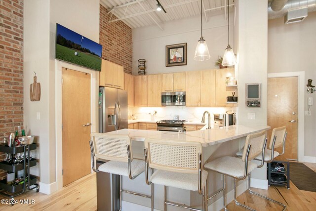 kitchen with appliances with stainless steel finishes, a towering ceiling, light brown cabinetry, kitchen peninsula, and light wood-type flooring