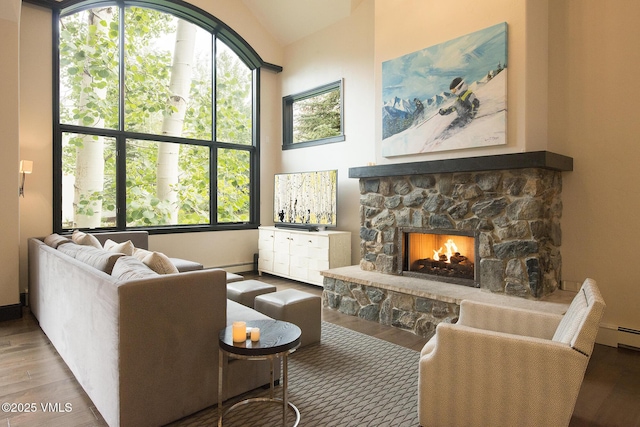 living room with baseboard heating, wood-type flooring, a stone fireplace, and a high ceiling