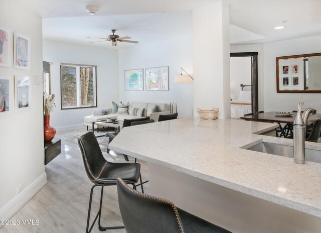 kitchen with sink, a kitchen breakfast bar, ceiling fan, light stone countertops, and light wood-type flooring