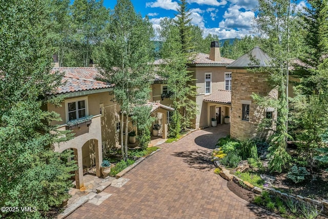 mediterranean / spanish-style home featuring a tile roof, stone siding, decorative driveway, stucco siding, and a chimney