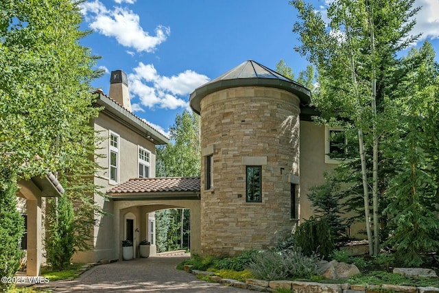 view of home's exterior with decorative driveway, a tile roof, a chimney, stucco siding, and stone siding