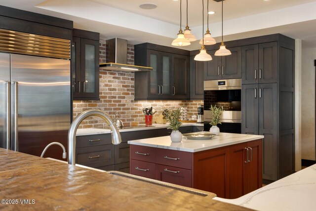 kitchen with glass insert cabinets, appliances with stainless steel finishes, light stone counters, wall chimney range hood, and pendant lighting