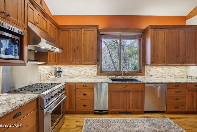 kitchen with appliances with stainless steel finishes, brown cabinets, light stone countertops, under cabinet range hood, and a sink