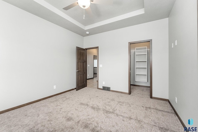 unfurnished bedroom with a raised ceiling, a closet, a walk in closet, ceiling fan, and light carpet