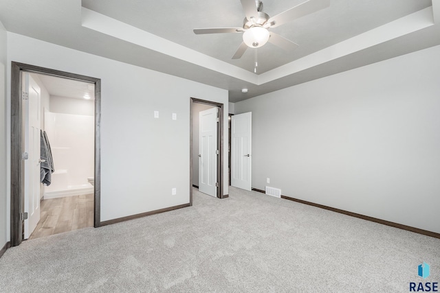 unfurnished bedroom with a raised ceiling, ceiling fan, light colored carpet, and ensuite bathroom