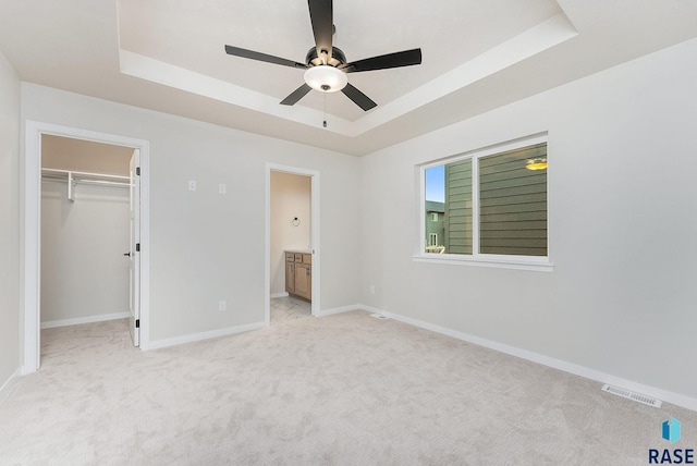 unfurnished bedroom with a raised ceiling, ensuite bathroom, a spacious closet, and ceiling fan