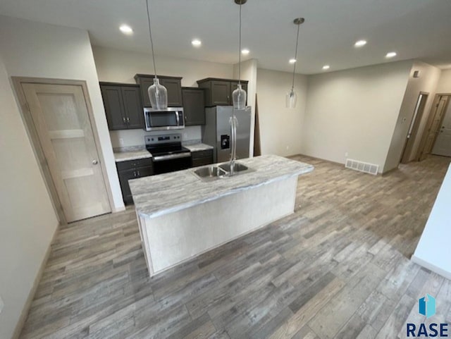 kitchen with a center island with sink, stainless steel appliances, hanging light fixtures, light stone counters, and wood-type flooring