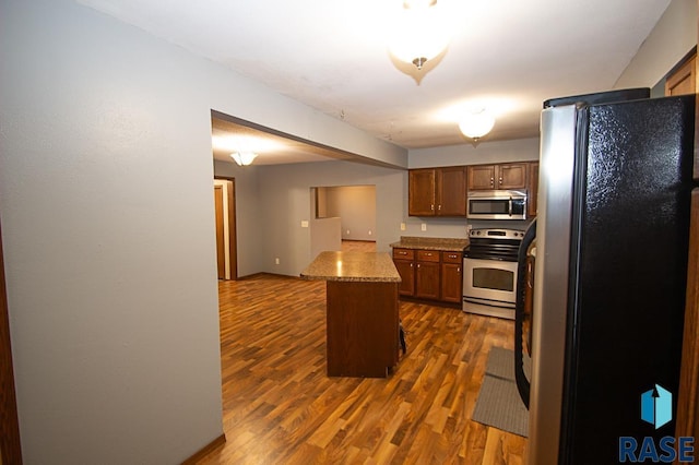 kitchen featuring brown cabinetry, appliances with stainless steel finishes, dark wood-style flooring, a center island, and light stone countertops