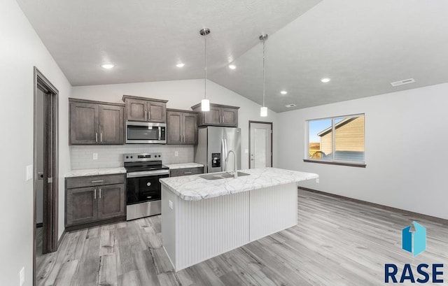 kitchen with light wood-type flooring, an island with sink, hanging light fixtures, stainless steel appliances, and vaulted ceiling