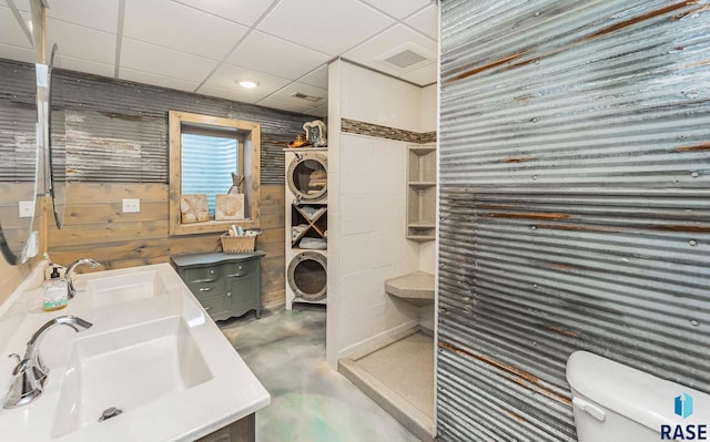 bathroom featuring toilet, stacked washer and dryer, wooden walls, vanity, and concrete flooring