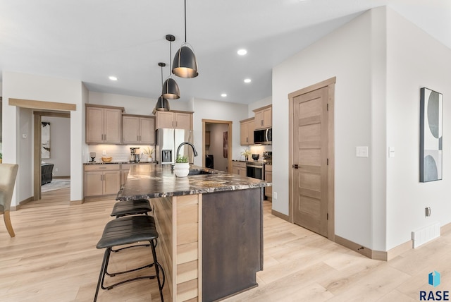 kitchen featuring dark stone countertops, pendant lighting, an island with sink, appliances with stainless steel finishes, and light hardwood / wood-style floors