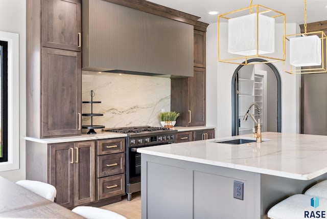 kitchen with light stone counters, sink, an island with sink, stainless steel stove, and backsplash