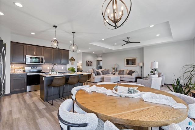 dining room with ceiling fan with notable chandelier, light wood-style flooring, recessed lighting, and a raised ceiling
