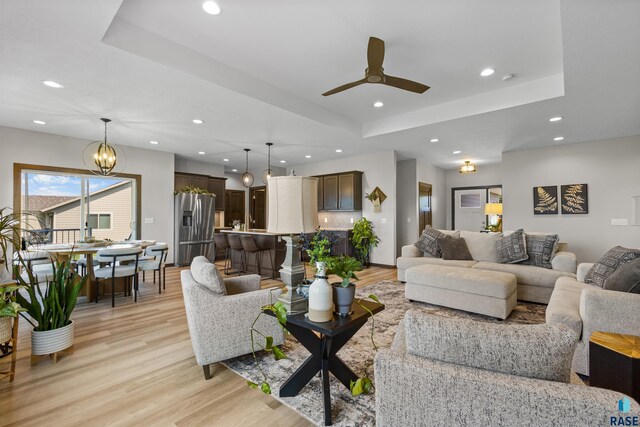 living area with ceiling fan with notable chandelier, a tray ceiling, light wood finished floors, and recessed lighting