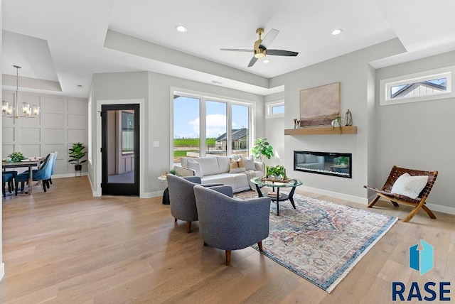 living room with ceiling fan with notable chandelier, light hardwood / wood-style floors, a tray ceiling, and a wealth of natural light