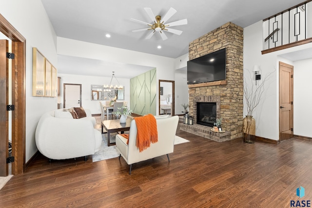 living room with ceiling fan with notable chandelier, a stone fireplace, and dark wood-type flooring