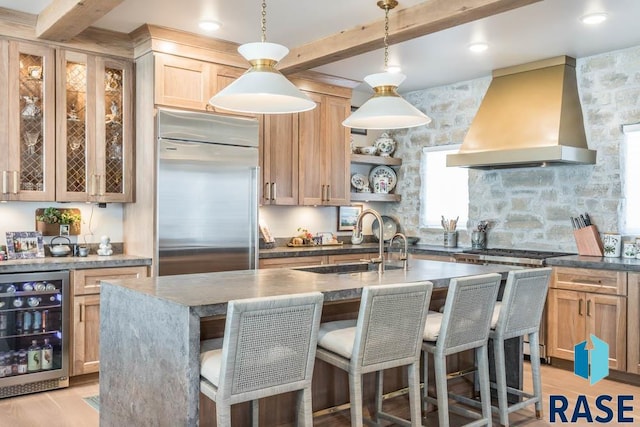 kitchen featuring an island with sink, wine cooler, stainless steel appliances, sink, and wall chimney range hood