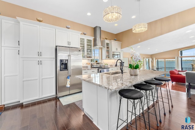 kitchen with an island with sink, sink, wall chimney range hood, white cabinetry, and appliances with stainless steel finishes