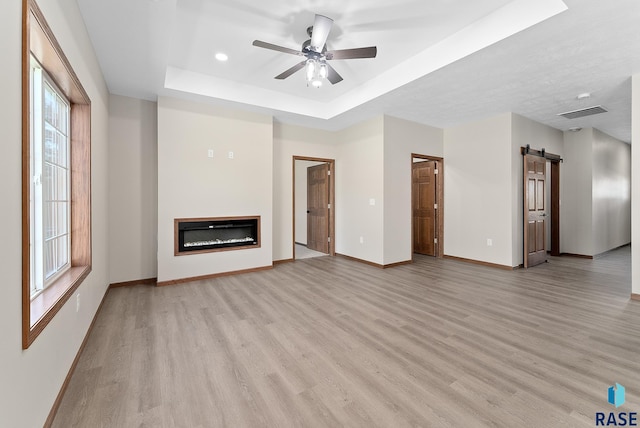 unfurnished living room with ceiling fan, light hardwood / wood-style floors, a barn door, and a tray ceiling