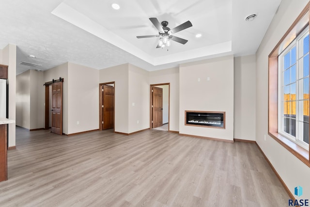 unfurnished living room with a barn door, a tray ceiling, ceiling fan, and light hardwood / wood-style flooring