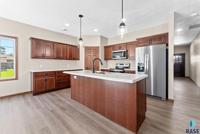 kitchen with hanging light fixtures, sink, a center island with sink, stainless steel appliances, and light wood-type flooring