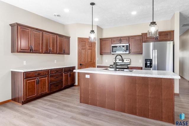 kitchen with an island with sink, stainless steel appliances, hanging light fixtures, and light wood-type flooring