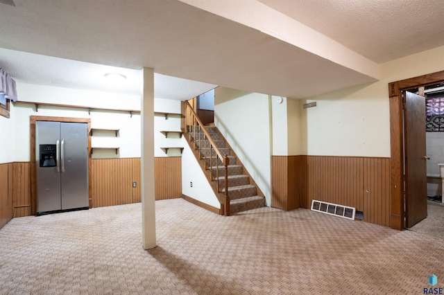 basement featuring a textured ceiling, carpet, wood walls, and stainless steel fridge with ice dispenser