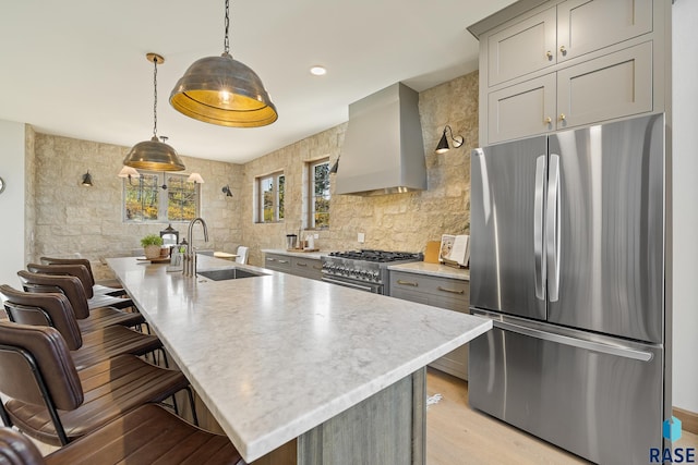 kitchen with wall chimney range hood, sink, hanging light fixtures, stainless steel appliances, and a kitchen island with sink