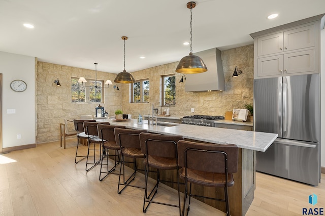 kitchen featuring a kitchen island with sink, wall chimney exhaust hood, stainless steel appliances, gray cabinets, and light stone counters