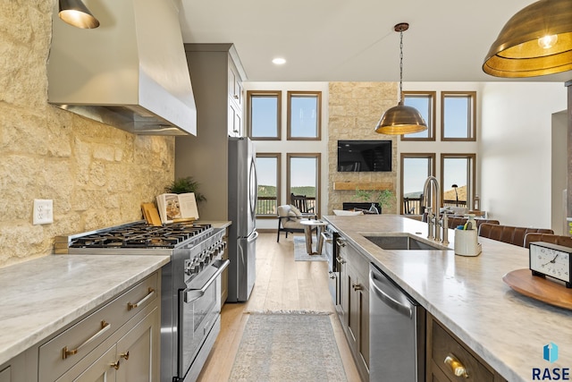 kitchen with wall chimney range hood, a stone fireplace, sink, appliances with stainless steel finishes, and light hardwood / wood-style floors