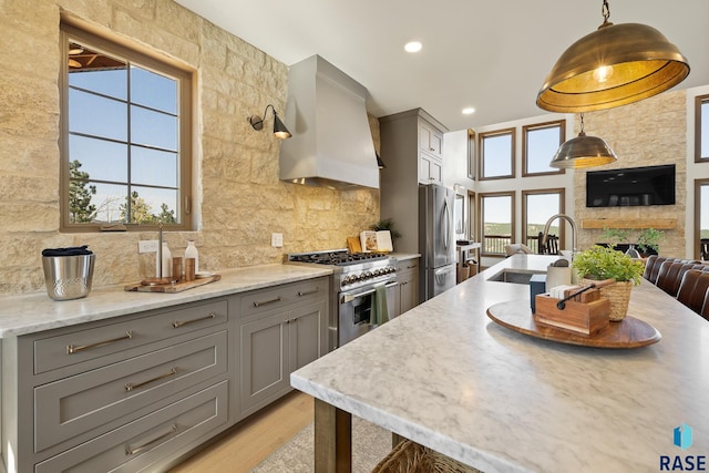 kitchen featuring wall chimney range hood, gray cabinetry, stainless steel appliances, light stone countertops, and decorative light fixtures