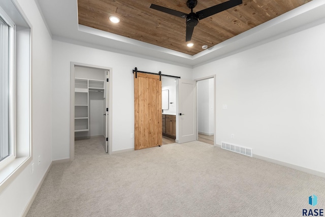 unfurnished bedroom with ceiling fan, a raised ceiling, a barn door, light colored carpet, and a walk in closet