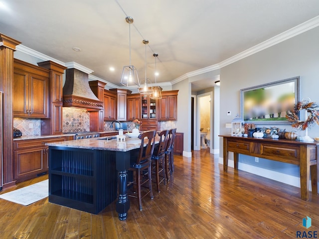 kitchen with an island with sink, premium range hood, ornamental molding, dark wood-type flooring, and light stone counters