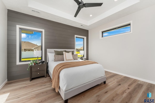 bedroom with ceiling fan, light hardwood / wood-style flooring, wooden walls, and a tray ceiling