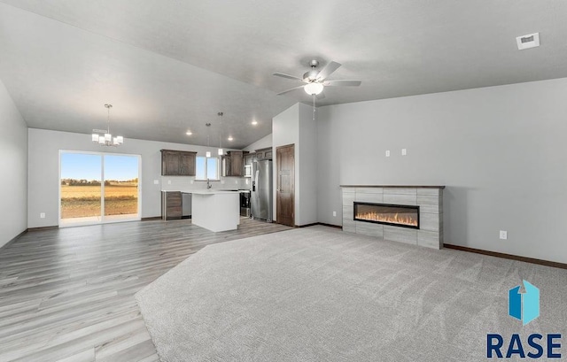 unfurnished living room with light wood-type flooring, a tiled fireplace, vaulted ceiling, and ceiling fan with notable chandelier