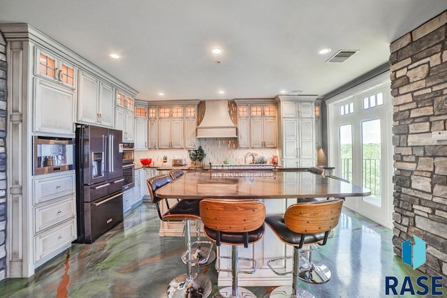 kitchen with stainless steel oven, high end black refrigerator, tasteful backsplash, an island with sink, and custom range hood