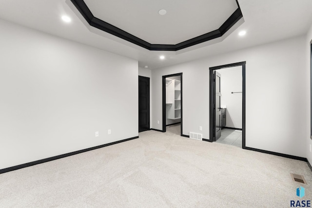 unfurnished bedroom with ensuite bathroom, crown molding, a tray ceiling, a walk in closet, and light carpet