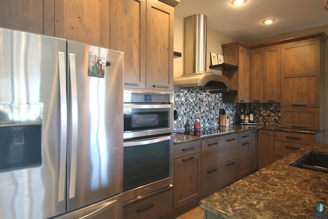 kitchen featuring brown cabinets, backsplash, appliances with stainless steel finishes, wall chimney range hood, and dark stone counters