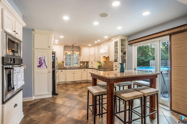 kitchen featuring appliances with stainless steel finishes, premium range hood, white cabinetry, hanging light fixtures, and a breakfast bar area