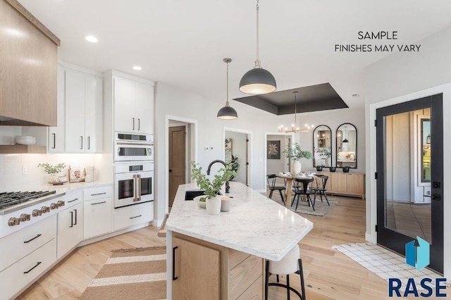 kitchen featuring white appliances, an island with sink, a tray ceiling, light wood-type flooring, and a sink