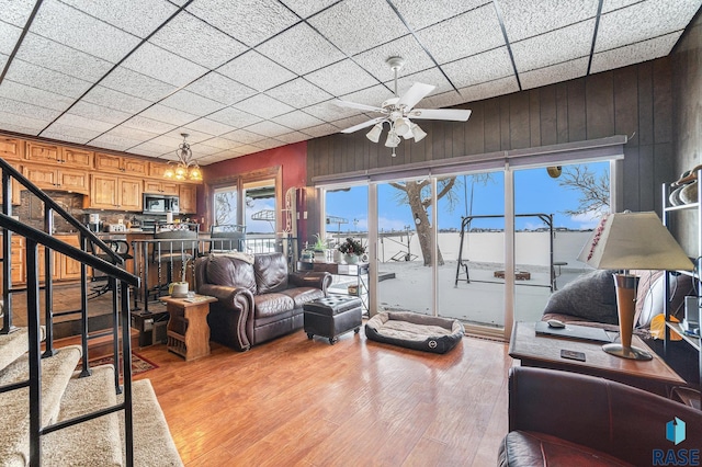 living room featuring ceiling fan with notable chandelier, light hardwood / wood-style floors, a drop ceiling, and wooden walls