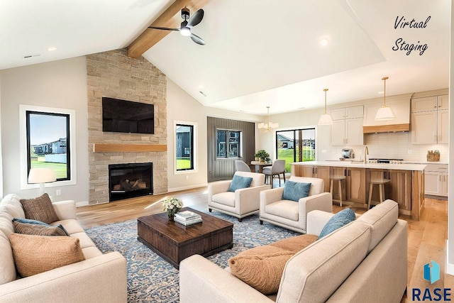 living room with lofted ceiling with beams, ceiling fan with notable chandelier, a stone fireplace, sink, and light hardwood / wood-style flooring